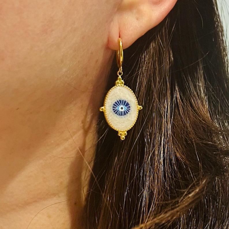 Op de afbeelding: Gouden oorbellen met een witte ovale hanger met een blauw boze oog ontwerp. De hanger is omgeven door een gouden rand en heeft een gouden ketting.