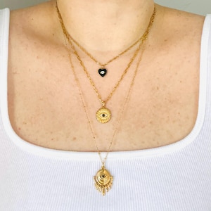May include: A layered gold-tone necklace set featuring three distinct chains. The shortest chain has a small black heart pendant with an eye motif. The middle chain holds a circular sunburst pendant with a central eye. The longest chain features a large, ornate eye-shaped pendant with radiating rays.