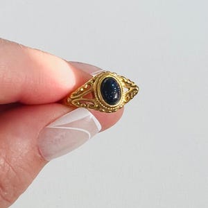 May include: A gold-colored ring with an oval, dark blue stone. The ring has an ornate, filigree design around the stone and on the band. The ring is held up against a white background.