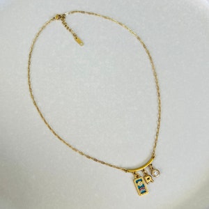 Peut inclure: Un collier en cha&icirc;ne dor&eacute;e avec un pendentif repr&eacute;sentant un soleil, une lettre 'R' et un petit strass transparent. Le pendentif est attach&eacute; &agrave; une barre dor&eacute;e.