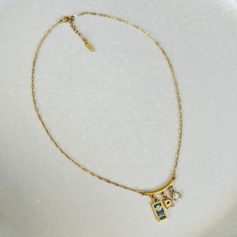 Peut inclure: Un collier en cha&icirc;ne dor&eacute;e avec un pendentif repr&eacute;sentant un soleil, une lettre 'R' et un petit strass transparent. Le pendentif est attach&eacute; &agrave; une barre dor&eacute;e.