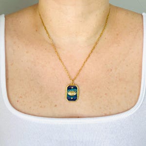May include: A gold-toned chain necklace featuring a rectangular pendant with a dark blue enamel background. The pendant displays a central eye motif with clear stones, surrounded by small gold star accents and a textured gold border frame.