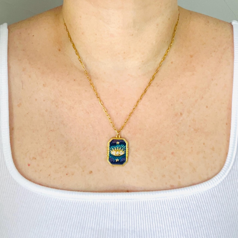 May include: A gold-toned chain necklace featuring a rectangular pendant with a dark blue enamel background. The pendant displays a central eye motif with clear stones, surrounded by small gold star accents and a textured gold border frame.