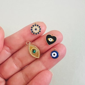 May include: Four gold-toned metal charms featuring eye motifs. One is a circular charm with blue and clear stones, one is a black heart with a central eye, one is an oval charm with a green stone, and one is a small circular blue and white eye.