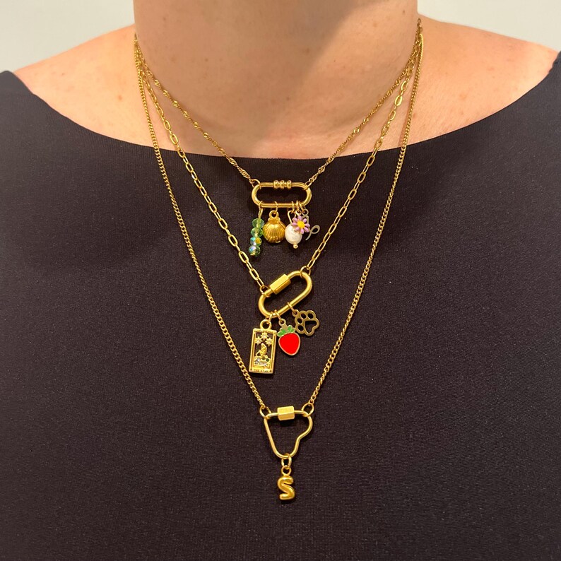 May include: A layered gold necklace with multiple charms, including a strawberry, a flower, a seashell, and a small green bead. The necklace is on a model wearing a black shirt.