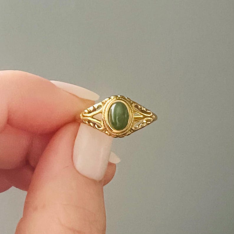 May include: A gold ring with an oval, green gemstone. The ring has an ornate, filigree design around the gemstone. The ring is held up against a grey background.