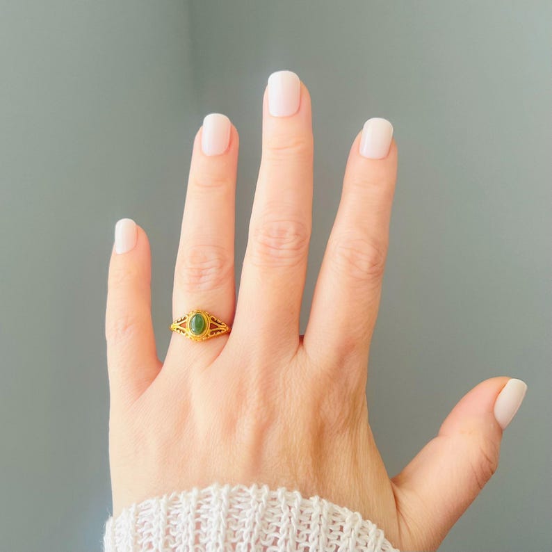 May include: A gold ring with an oval green gemstone is worn on a finger. The ring features an ornate filigree design around the gemstone. The hand is against a grey background, and the person is wearing a white sweater.