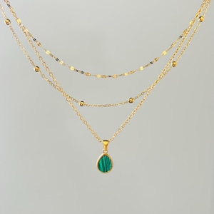 May include: A layered gold necklace with three strands. The bottom strand features a teardrop-shaped pendant with a green stone. The other strands have small gold beads and flat gold pieces. The necklace is displayed against a light gray background.