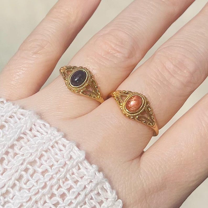 May include: Two gold rings with oval-shaped gemstones. One ring features a dark blue gemstone, and the other has a reddish-orange stone. Both rings have intricate filigree detailing around the gemstones and on the bands. The rings are displayed on a person's fingers.