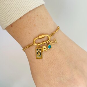 May include: Gold charm bracelet with a carabiner clasp, featuring a small blue gemstone, a letter 'R' charm, a zodiac symbol charm, and a rectangular charm with a black and white design and rhinestones.