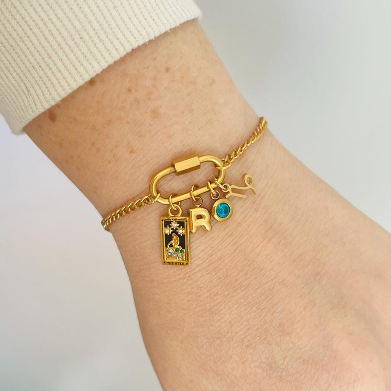 May include: Gold charm bracelet with a carabiner clasp, featuring a small blue gemstone, a letter 'R' charm, a zodiac symbol charm, and a rectangular charm with a black and white design and rhinestones.