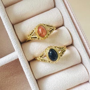 May include: Two gold-colored rings with oval gemstones, one orange and one dark blue, rest in a pink jewelry box. The rings have ornate filigree detailing around the gemstones. The jewelry box has a soft, padded interior.