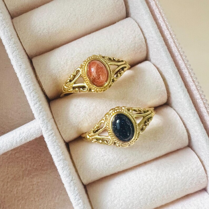 May include: Two gold-colored rings with oval gemstones, one orange and one dark blue, rest in a pink jewelry box. The rings have ornate filigree detailing around the gemstones. The jewelry box has a soft, padded interior.