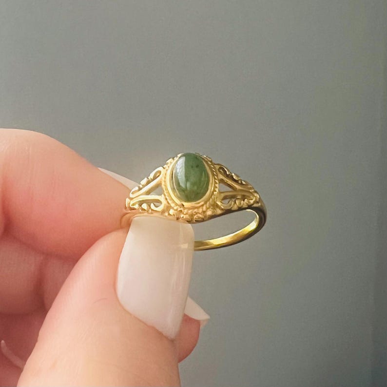 May include: A gold ring with an oval, green gemstone. The ring has an ornate, filigree design around the gemstone and along the band. The ring is held up against a grey background.