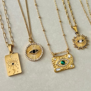 May include: Four gold-toned necklaces featuring various pendants: a rectangular tag with an eye and moon, a circular sunburst with a central eye, a textured rectangular plate with a green stone eye, and a sunburst charm with a central clear stone.