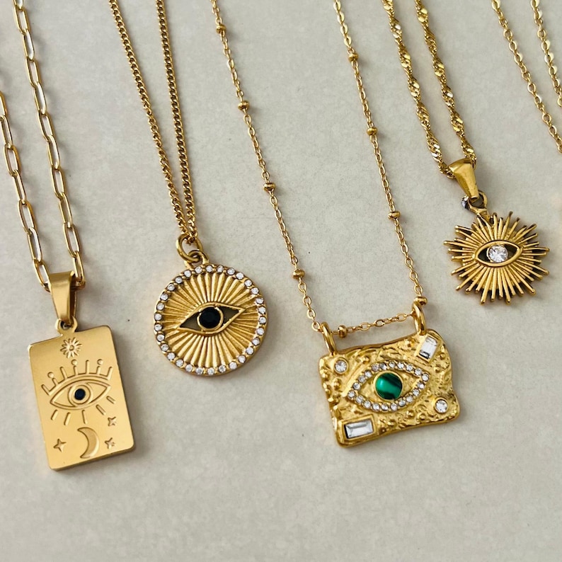 May include: Four gold-toned necklaces featuring various pendants: a rectangular tag with an eye and moon, a circular sunburst with a central eye, a textured rectangular plate with a green stone eye, and a sunburst charm with a central clear stone.