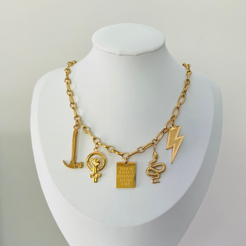 May include: Gold chain necklace with multiple charms, including a hammer, a fist, a snake, a lightning bolt, and a square charm with the text "This is what a feminist looks like".