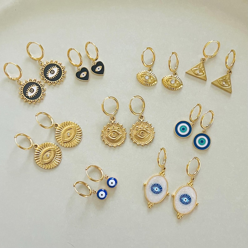 Op de afbeelding: Een verzameling gouden creolen met verschillende bedels, waaronder boze ogen, harten en driehoeken. De bedels zijn versierd met blauw en wit emaille en strass.