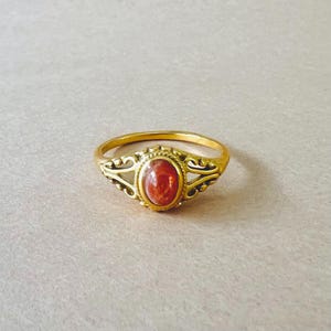May include: A gold-colored ring with an oval, reddish-orange stone. The ring has an ornate design around the stone and on the band's shoulders. The ring is set against a neutral background.