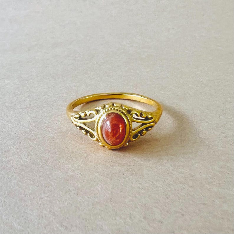 May include: A gold-colored ring with an oval, reddish-orange stone. The ring has an ornate design around the stone and on the band's shoulders. The ring is set against a neutral background.