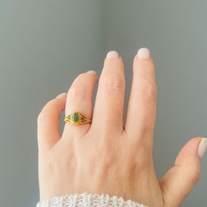 May include: A gold ring with an oval, green gemstone is shown on a hand. The ring features an intricate, filigree pattern around the gemstone and on the band. The background is a plain, light grey colour.