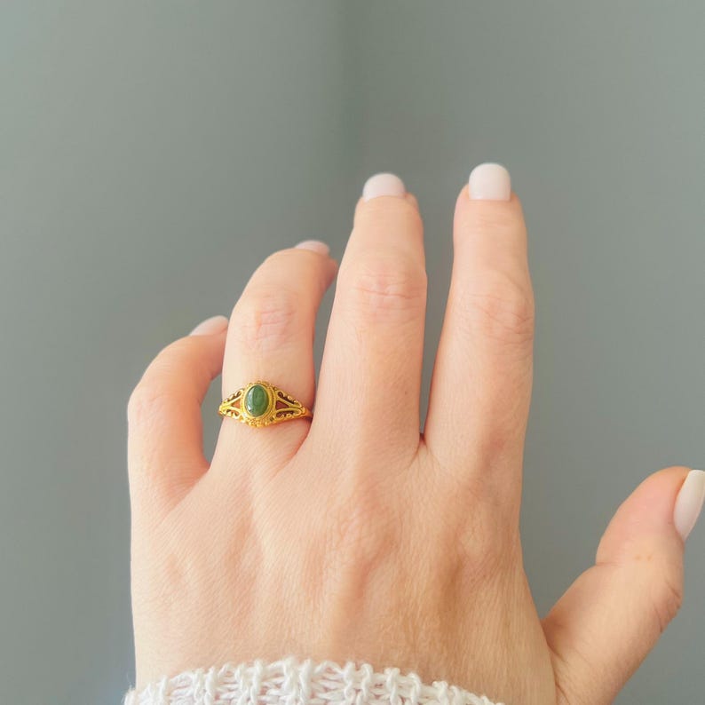 May include: A gold ring with an oval, green gemstone is shown on a hand. The ring features an intricate, filigree pattern around the gemstone and on the band. The background is a plain, light grey colour.