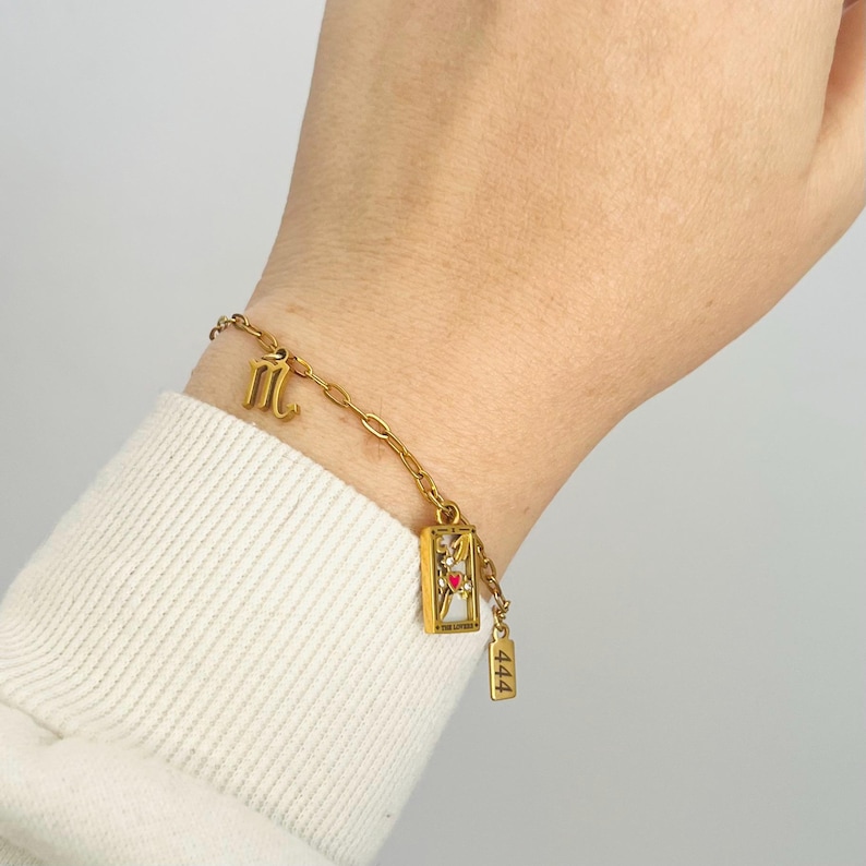 May include: Gold chain bracelet with a charm that says "444" and a charm with a small red heart inside a gold frame. The bracelet also has a charm with the astrological symbol for Scorpio.