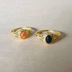 May include: Two gold-colored rings with oval gemstones. One ring features a reddish-orange stone, the other a dark blue stone. Both rings have an ornate, filigree design around the gemstone setting.