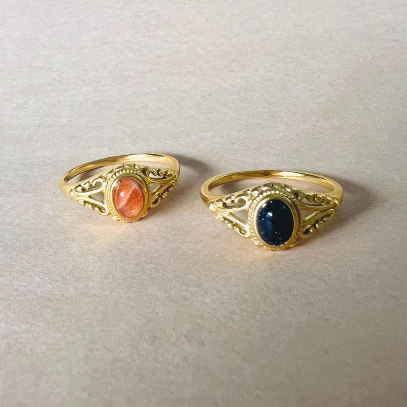 May include: Two gold-colored rings with oval gemstones. One ring features a reddish-orange stone, the other a dark blue stone. Both rings have an ornate, filigree design around the gemstone setting.