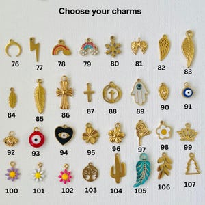 May include: A selection of gold-toned charms with various designs, including a crescent moon, lightning bolt, rainbow, snowflake, wings, feather, cross, hamsa hand, evil eye, heart, flower, cactus, and tree. The charms are numbered 76 through 107.