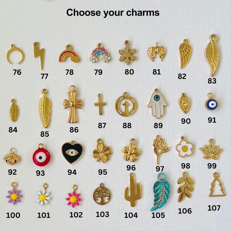 May include: A selection of gold-toned charms with various designs, including a crescent moon, lightning bolt, rainbow, snowflake, wings, feather, cross, hamsa hand, evil eye, heart, flower, cactus, and tree. The charms are numbered 76 through 107.