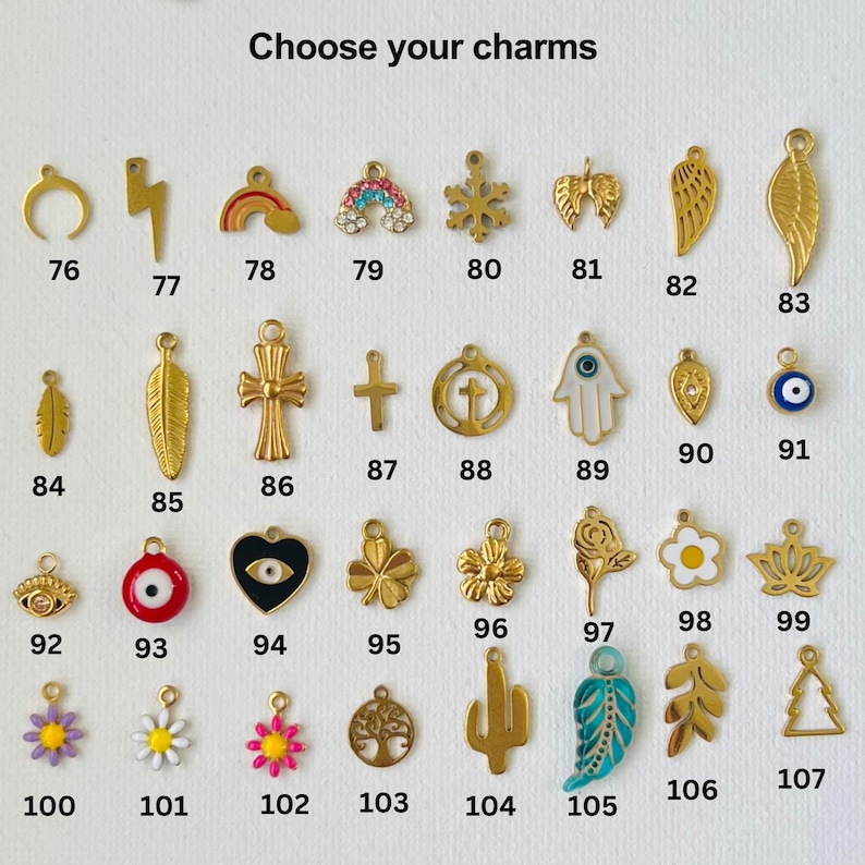 May include: A selection of gold-toned charms with various designs, including a crescent moon, lightning bolt, rainbow, snowflake, wings, feather, cross, hamsa hand, evil eye, heart, flower, cactus, and tree. The charms are numbered 76 through 107.