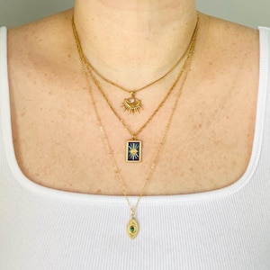 May include: A layered gold necklace set consisting of three chains. The top chain features a sunburst pendant with a clear stone. The middle chain holds a rectangular blue enamel pendant with a gold starburst. The bottom chain has an oval pendant.