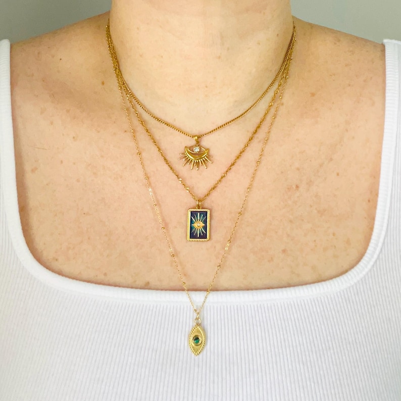 May include: A layered gold necklace set consisting of three chains. The top chain features a sunburst pendant with a clear stone. The middle chain holds a rectangular blue enamel pendant with a gold starburst. The bottom chain has an oval pendant.
