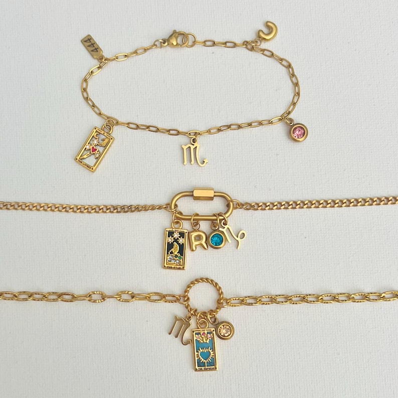 May include: Three gold chain bracelets with charms. The top bracelet has a pink crystal, a small gold charm with the letter "M" and a tarot card charm. The middle bracelet has a gold clasp, a gold charm with the letter "R", a blue crystal, a gold charm with the letter "O", and a tarot card charm. The bottom bracelet has a gold circle charm, a gold charm with the letter "M", and a tarot card charm.