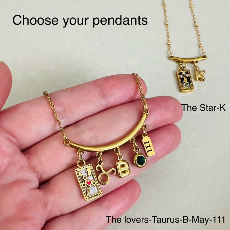 Peut inclure: Un collier en or avec une barre incurv&eacute;e et plusieurs breloques, dont une carte de tarot, un symbole du zodiaque, une lettre 'B', une pierre pr&eacute;cieuse verte et un breloque avec le num&eacute;ro '111'. Le texte 'The lovers-Taurus-B-May-111' est visible sous le collier.