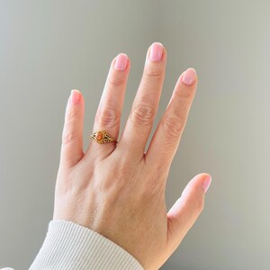 May include: A gold-colored ring with an oval, orange-colored stone. The ring has an ornate, filigree design around the stone and on the band. The ring is displayed on a hand with pink nail polish.
