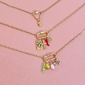 May include: Three gold necklaces with charms. The top necklace has a heart-shaped charm with the letter "S" on it. The middle necklace has a charm with a cross, a red chili pepper, and a paw print. The bottom necklace has a charm with a seashell, a pearl, a green bead, and a flower.