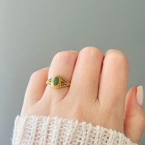 May include: A gold ring featuring an oval green gemstone. The ring has an intricate design with cut-out patterns. The ring is on a finger, with a white sweater visible in the background.