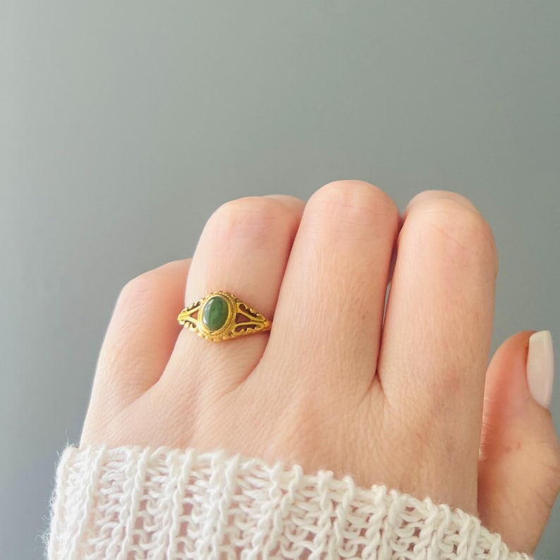 May include: A gold ring featuring an oval green gemstone. The ring has an intricate design with cut-out patterns. The ring is on a finger, with a white sweater visible in the background.