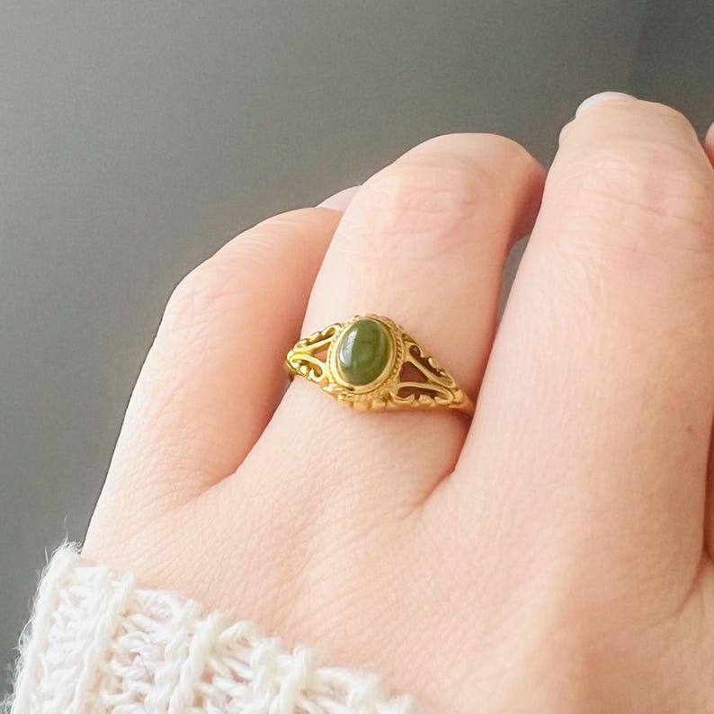 May include: A gold ring with an oval, olive-green gemstone. The ring has an ornate, filigree design around the stone and along the band. The ring is displayed on a hand with a white, textured sleeve.