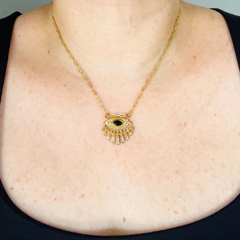 May include: A gold-toned necklace featuring a pendant shaped like an eye with a central black marquise-cut stone. The pendant has radiating gold rays extending downward and is suspended from a delicate gold-toned chain with oval links.