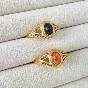 May include: Two gold-colored rings with oval gemstones. One ring features a dark blue stone with glitter-like inclusions, while the other has a reddish-orange stone. Both rings have ornate, filigree-style settings.