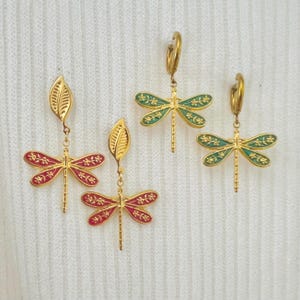 May include: Gold-toned dragonfly earrings with red and green enamel wings. The earrings feature intricate floral detailing and are suspended from gold leaf-shaped accents and hoop closures. The dragonfly design adds a touch of nature-inspired elegance.