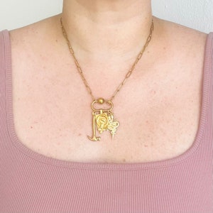 May include: A gold chain necklace with a variety of charms, including a hammer, a snake, a butterfly, and a small square charm with a design. The necklace is worn by a person with a light pink top.