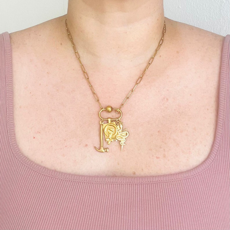 May include: A gold chain necklace with a variety of charms, including a hammer, a snake, a butterfly, and a small square charm with a design. The necklace is worn by a person with a light pink top.