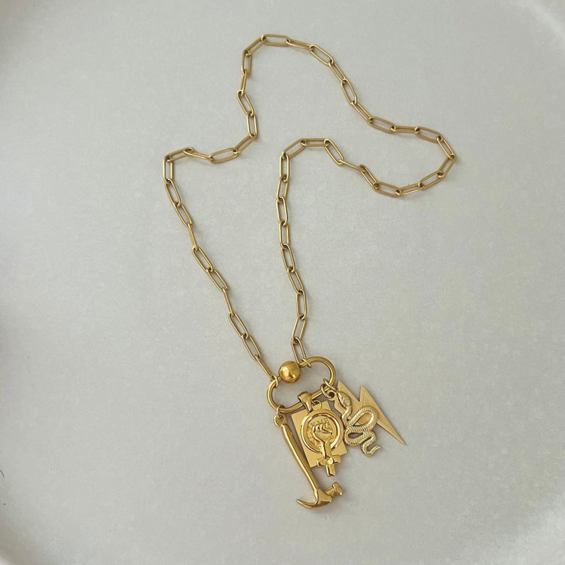 May include: A gold chain necklace with a charm featuring a hammer, a snake, a lightning bolt, and a fist. The chain is made of small, linked circles.