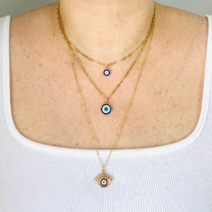 May include: A layered gold-tone necklace set featuring three chains of varying lengths. The shortest chain has a small circular evil eye charm, the middle chain holds a larger circular evil eye pendant, and the longest chain features an eye-shaped charm.