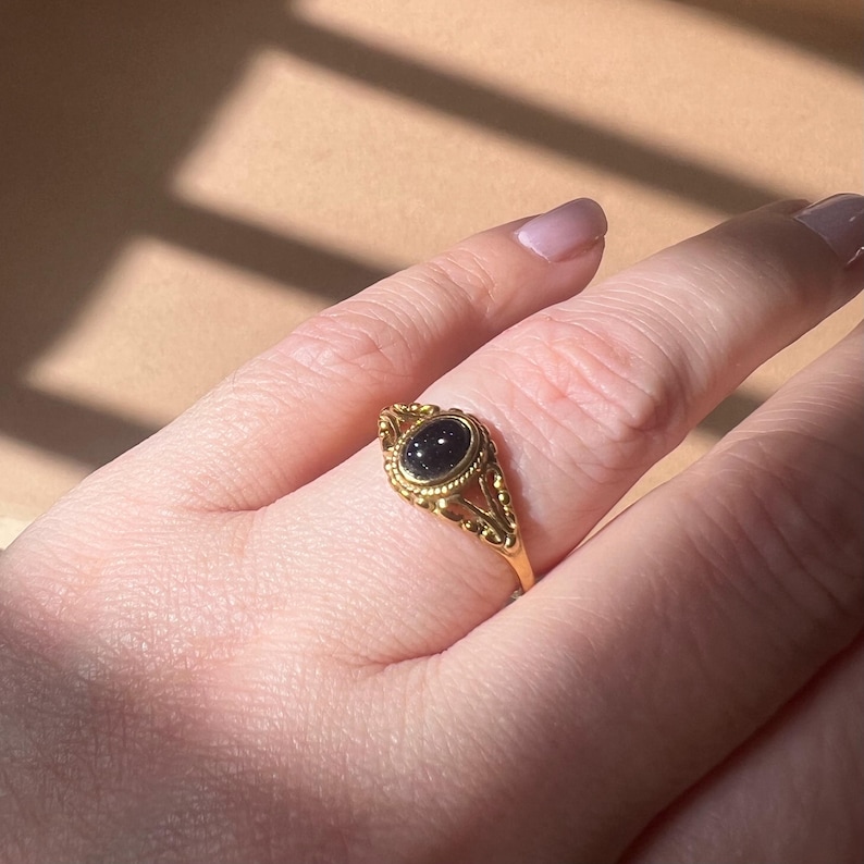 May include: A gold-colored ring with an oval, dark-colored stone. The ring has an ornate, filigree design around the stone and along the band. The ring is on a finger, with the background out of focus.
