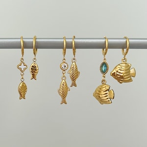 May include: A collection of gold-colored hoop earrings. Each earring features a unique charm, including fish, a four-leaf clover, and gemstones. The earrings are displayed on a silver bar against a plain background.
